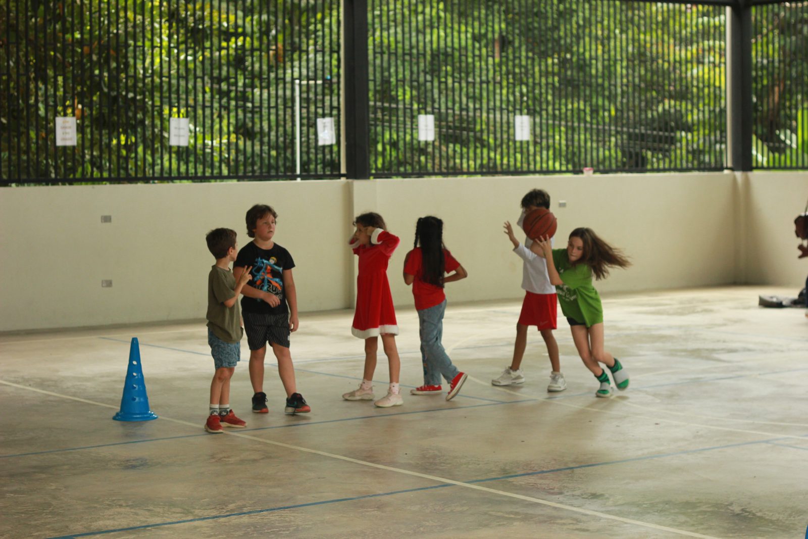 Six children playing basketball in a covered court beneath green fence backdrop