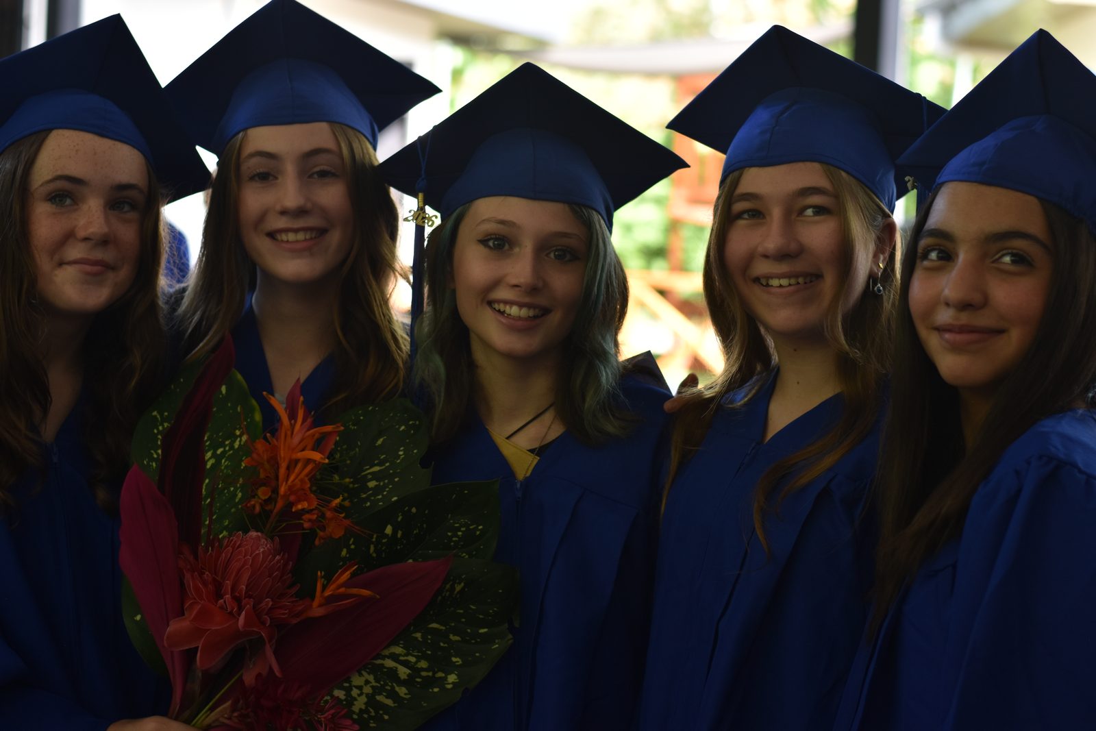 Five graduating students in blue caps and gowns hold flowers and smile at ceremony