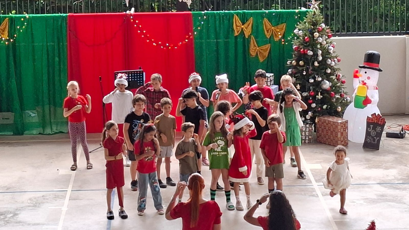 Young students perform on gymnasium floor with holiday backdrop and decorated Christmas tree