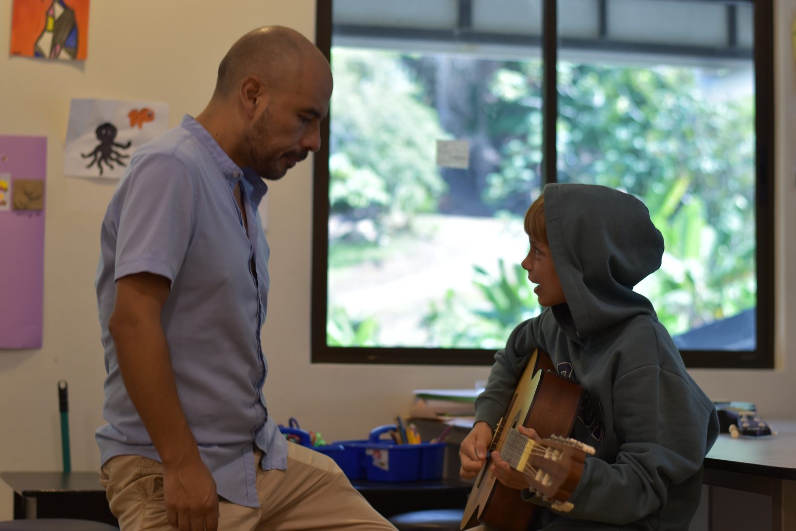 Teacher instructs student playing acoustic guitar in bright classroom overlooking landscape