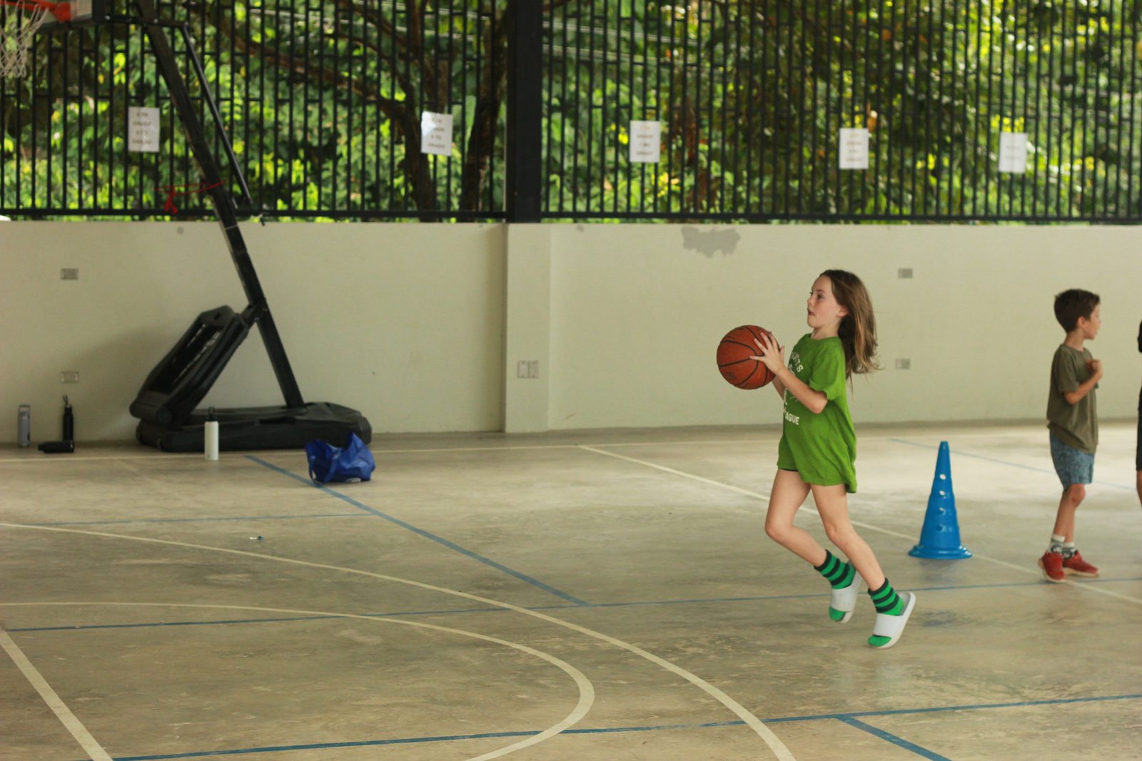 Student in green jersey runs with basketball on gymnasium court overlooking jungle
